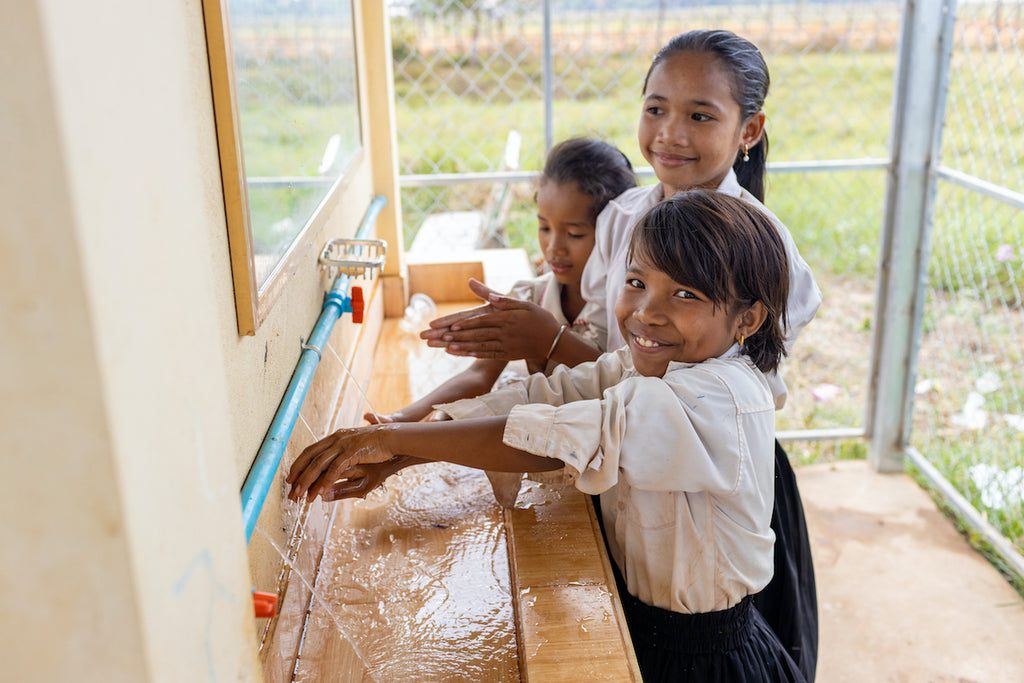 Three young girls in school uniforms smile while washing their hands at an outdoor sink with running water, near a wire fence and grassy area.