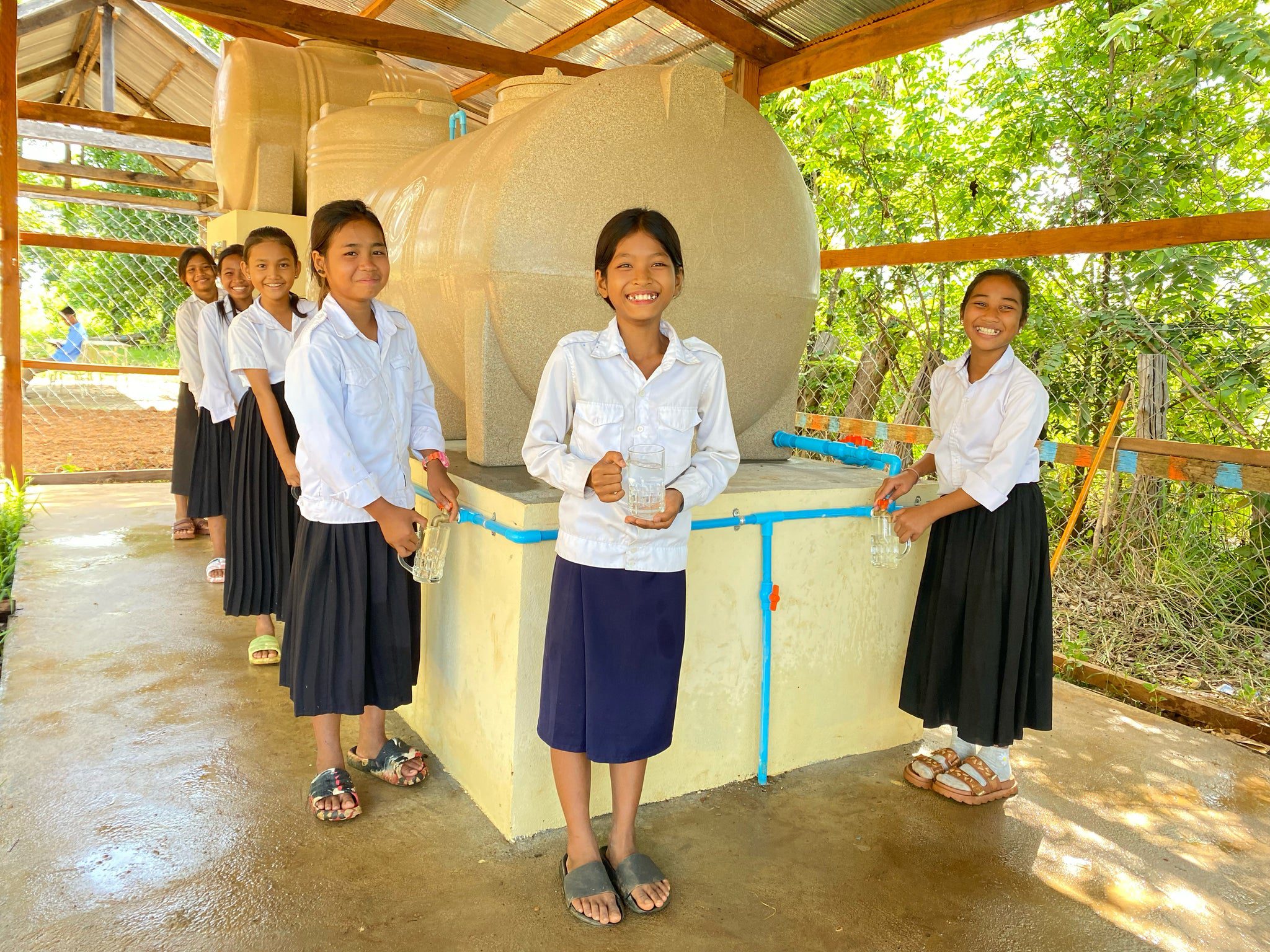 Seven smiling schoolgirls in uniform stand around a large water tank under a shelter, each holding a cup or bottle, with green trees and sunlight in the background.