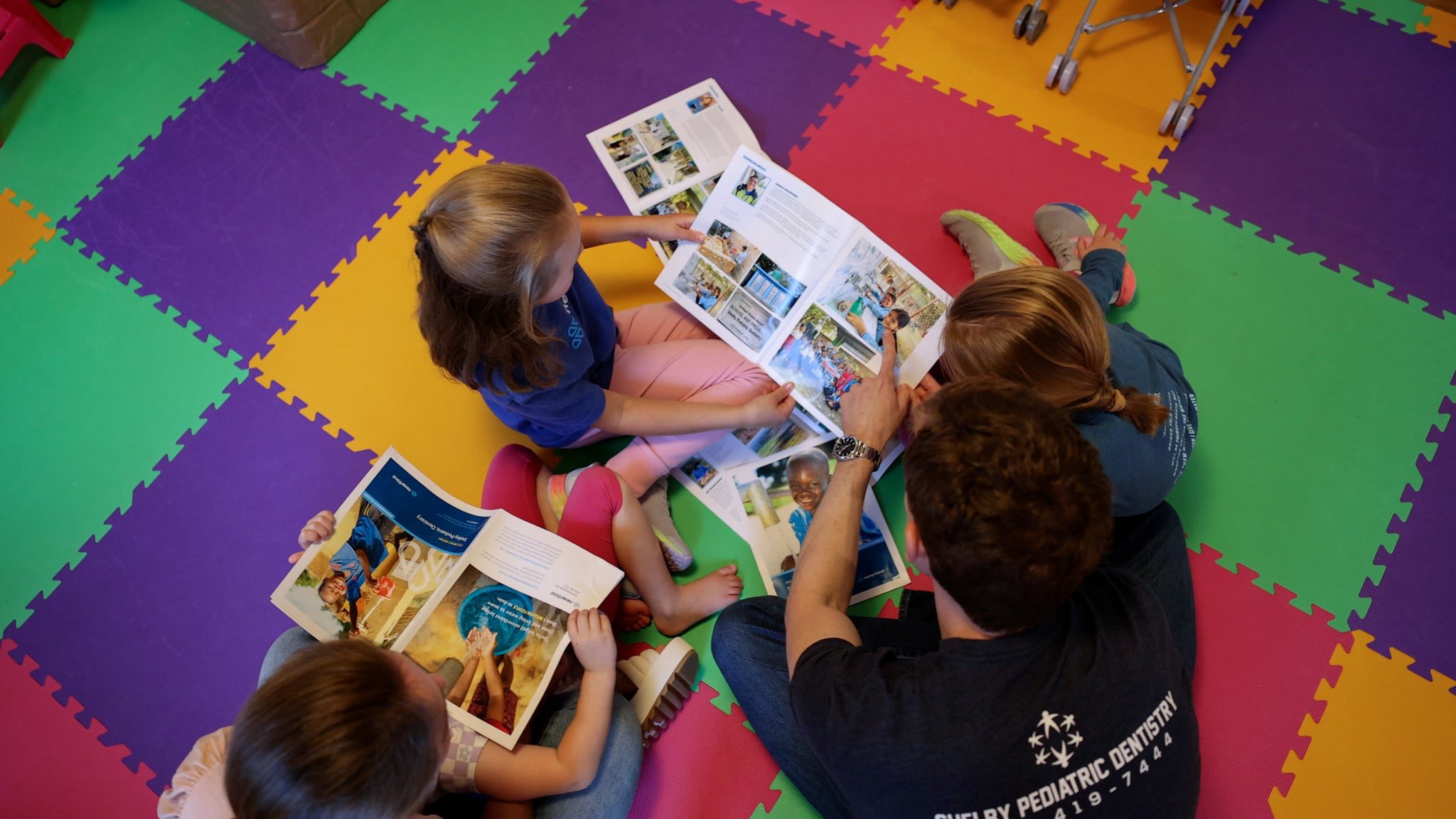 Four children sit in a circle on a colorful foam mat, looking at picture books together. The scene is bright and playful, suggesting a fun and educational group activity.