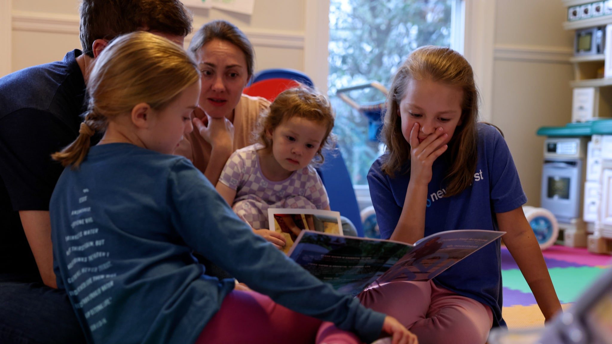 A group of children sit on the floor reading a picture book together, with an adult nearby. The children look engaged, and one girl covers her mouth in surprise or excitement. Colorful play mats are in the background.
