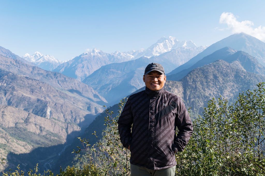 A man in a black jacket and cap stands smiling in front of a scenic mountain landscape with clear blue skies and snow-capped peaks in the background. Green bushes are visible in the foreground.