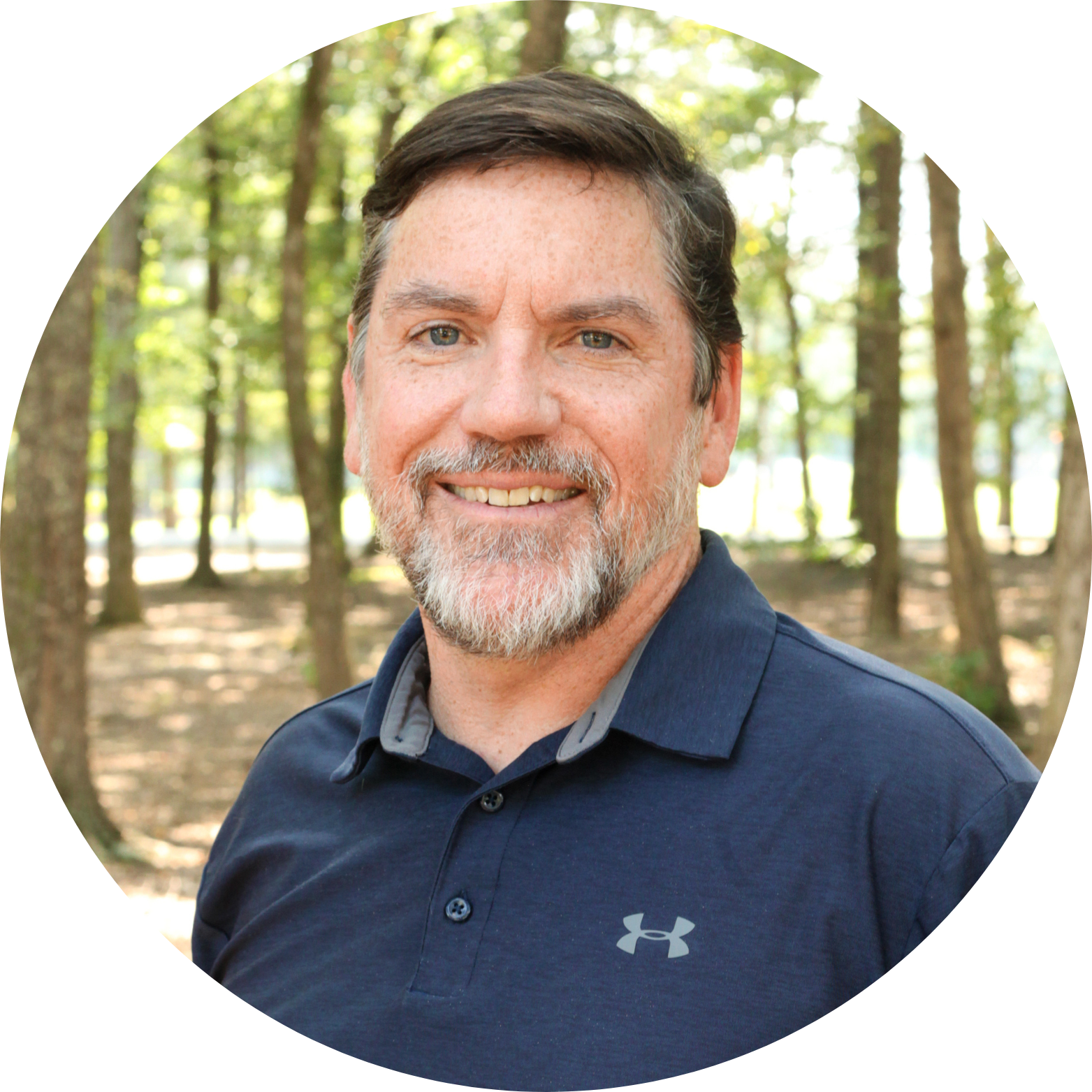 A man with short brown hair and a beard smiles at the camera. He is wearing a navy blue Under Armour polo shirt and standing in a sunlit forest with tall trees in the background.