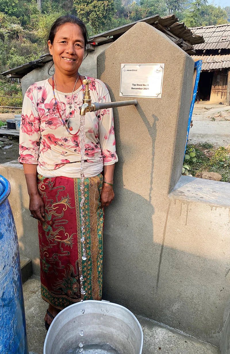 A woman stands smiling next to a tap with running water, filling a bucket. She is wearing a patterned skirt and light-colored shirt. Behind her is a stone structure with a plaque and a rural house in the background.