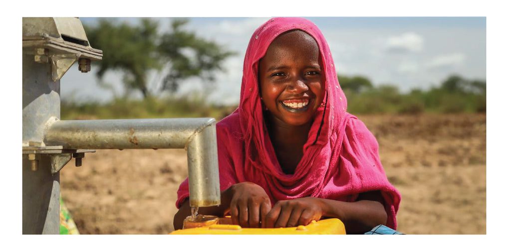 A smiling girl in a bright pink headscarf collects water from a hand pump into a yellow container, with a dry, rural landscape and trees in the background.