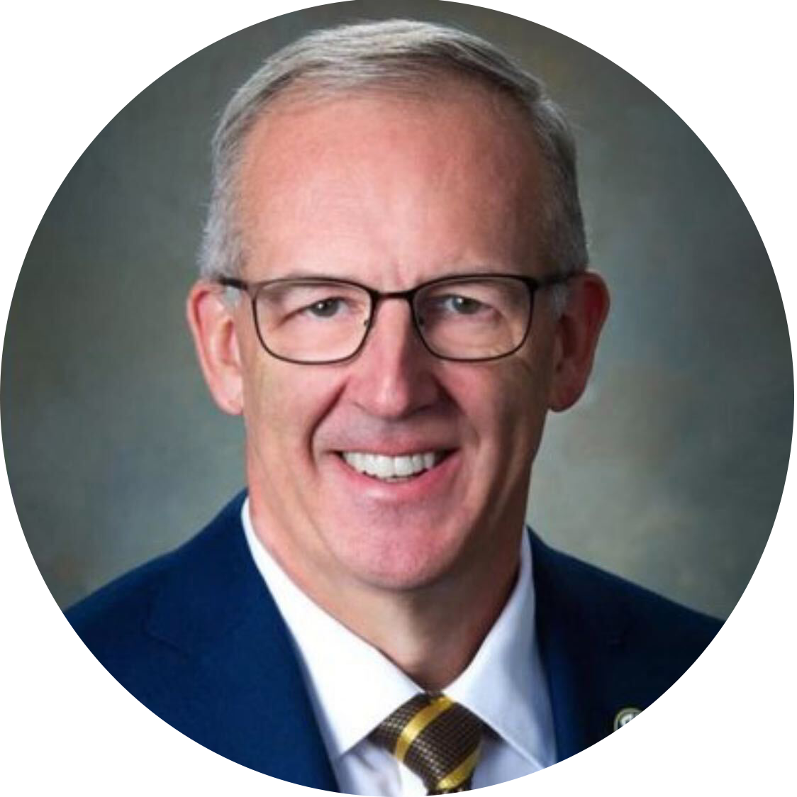 A man with short brown hair smiles at the camera, wearing a gray jacket. Green leaves and plants form a blurred natural background.