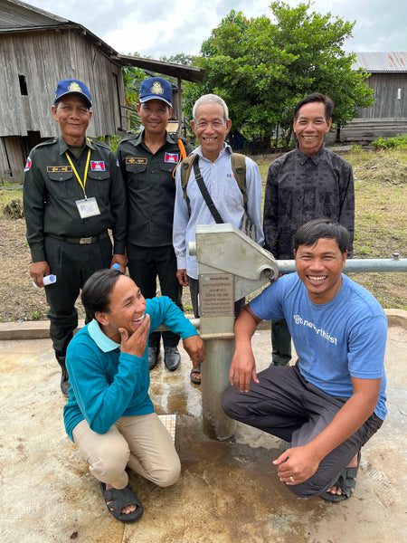 Six smiling men gather around a hand-powered water pump outdoors, with wooden buildings and greenery in the background. Two wear dark uniforms and hats, while the others are dressed casually.