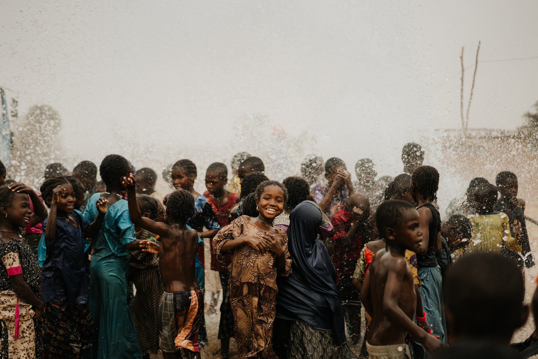 A group of children smile and play joyfully in water spray outdoors, with one girl in the center looking at the camera. The scene is lively and filled with splashes, and the kids wear colorful clothing.