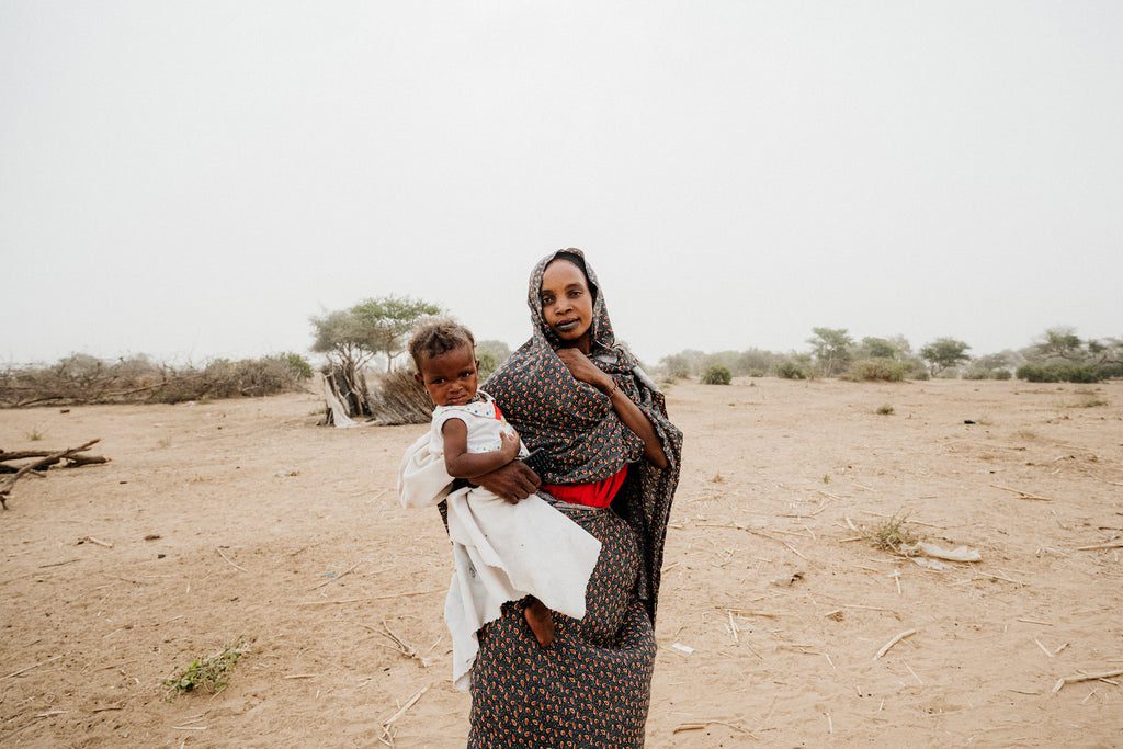 A woman in a patterned dress stands in a dry, barren landscape, holding a young child wrapped in a white cloth. Sparse trees and cloudy sky are visible in the background.