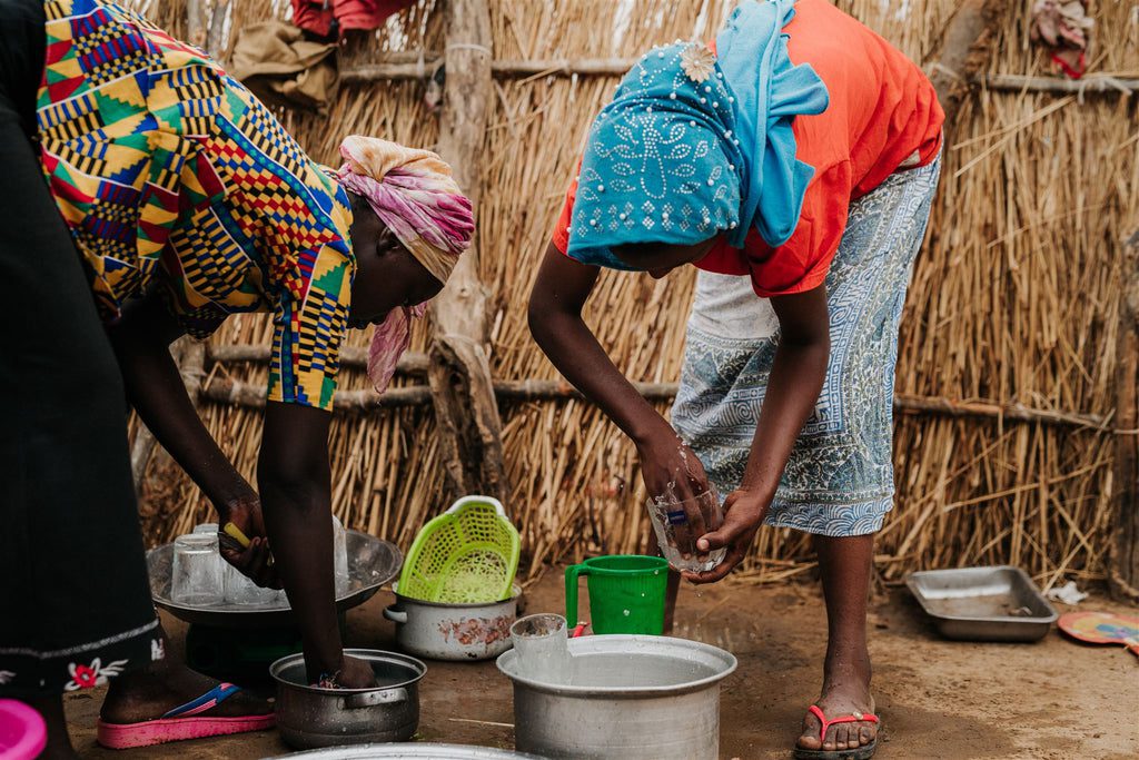 Two women wearing colorful clothing and headscarves bend over large metal bowls, washing dishes outdoors in front of a wall made of vertical sticks. Various kitchen items are scattered around them.