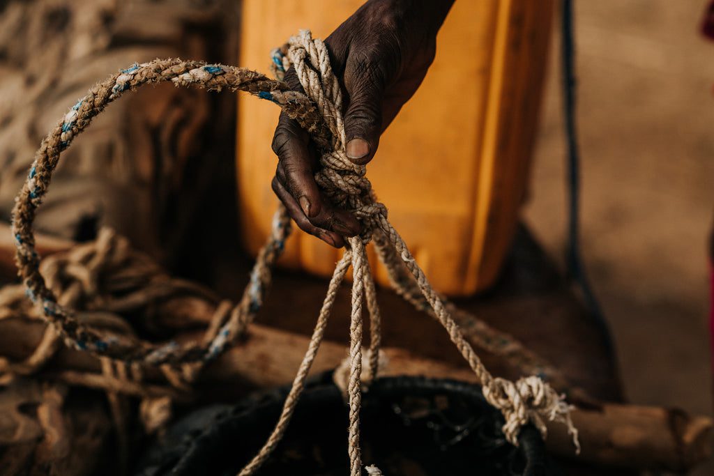 A close-up of a person’s hand holding and tying thick, weathered ropes, with a yellow plastic container and blurred objects in the background. The scene suggests manual labor or preparation for work.