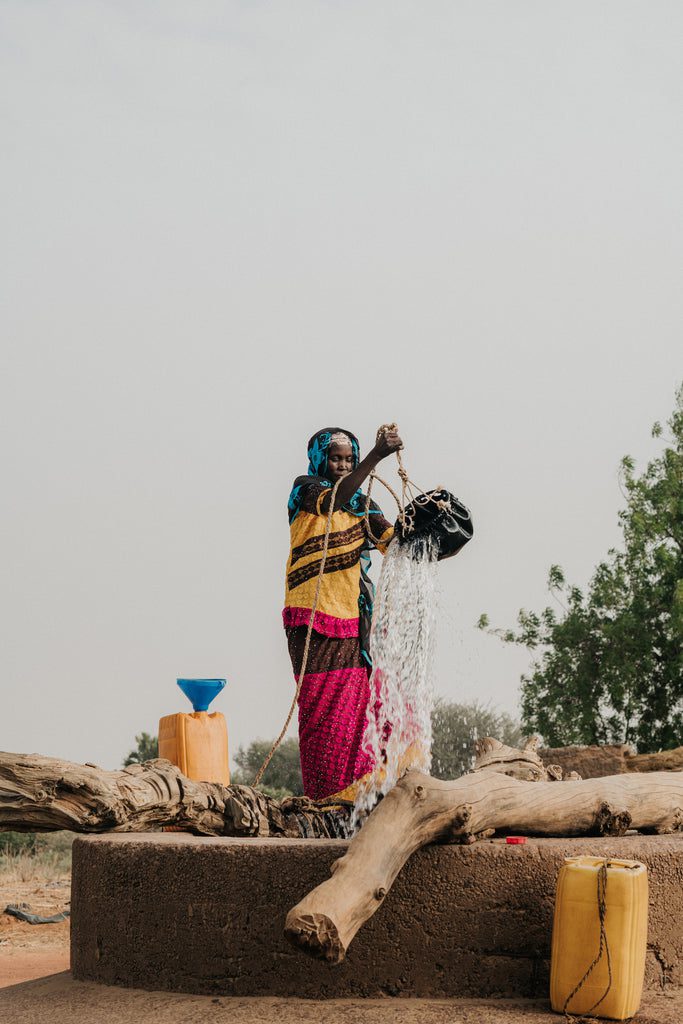 A woman in colorful clothing pours water from a bucket at a well in a rural outdoor setting, with yellow and blue containers nearby and trees in the background.