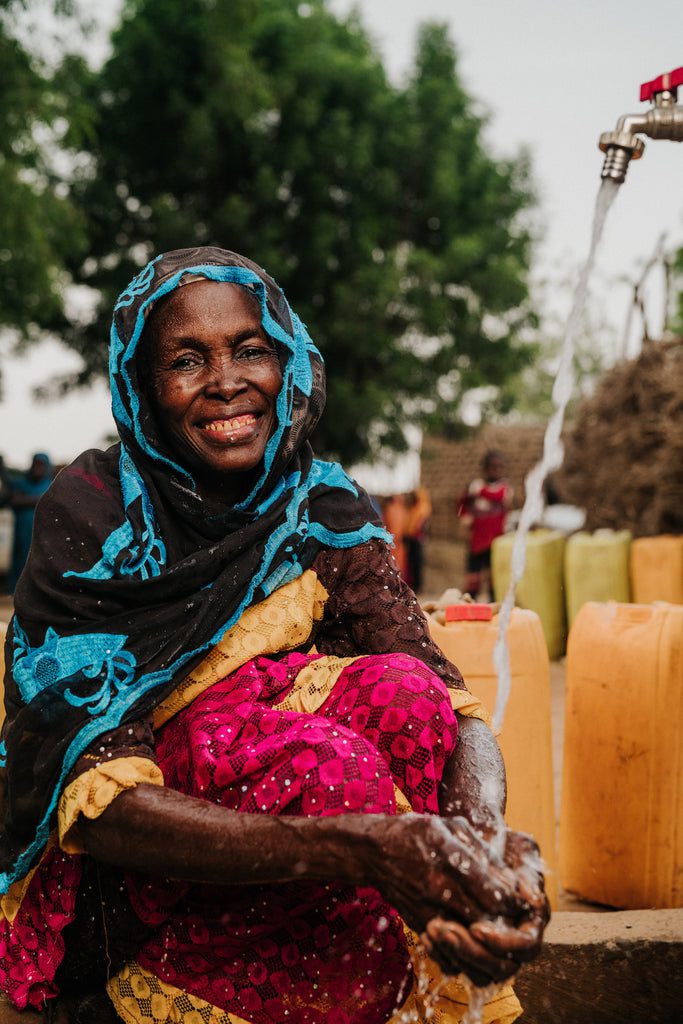 An older woman wearing colorful clothing and a headscarf smiles while washing her hands under running water from a tap, with yellow water containers and trees in the background.