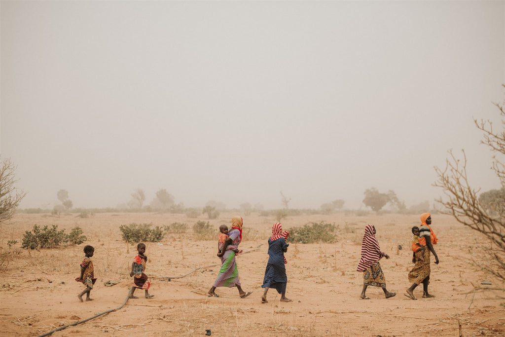 A group of women and children walk in single file across a dry, dusty landscape with sparse vegetation, dressed in colorful clothing and headscarves. The sky appears hazy, and the environment looks arid.