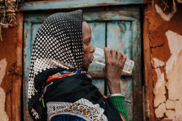 A woman wearing a patterned headscarf and colorful clothing drinks water from a glass while standing in front of a weathered, blue wooden door.