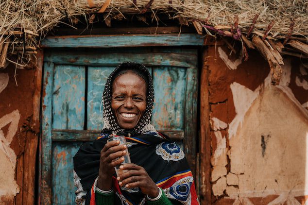 A smiling woman stands in front of a rustic, blue wooden door with a thatched roof above. She is wearing a colorful patterned shawl and holds a cylindrical object in her hands.