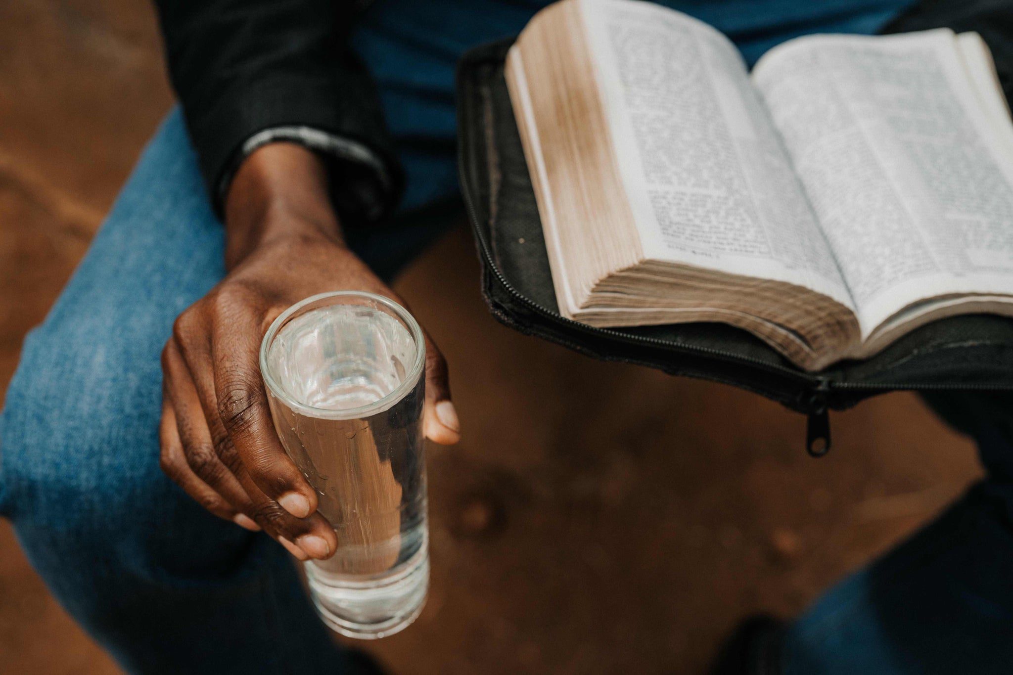 A person wearing blue jeans holds a clear glass of water in one hand and an open book with text in the other hand, seated on a brown surface.
