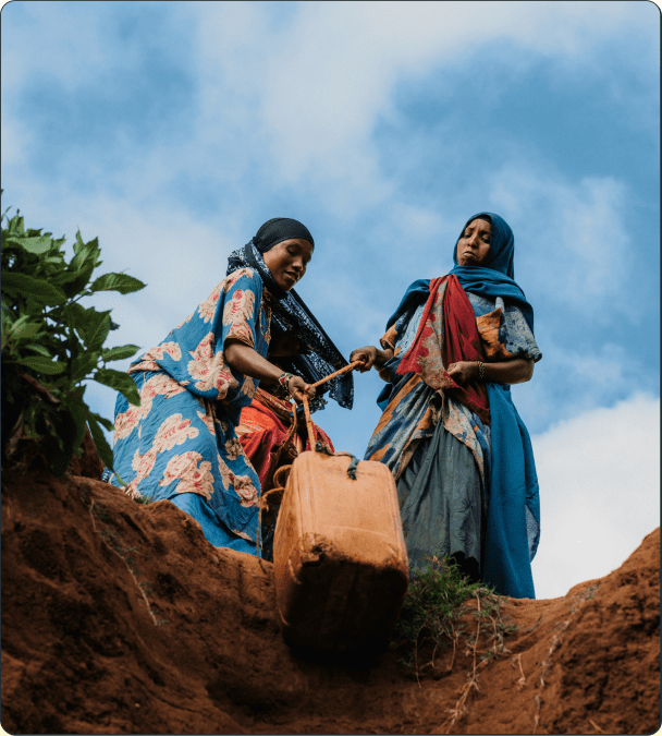 Two women in colorful clothing stand on a dirt slope, working together to pull up a large container with a rope. The sky is blue with clouds in the background.