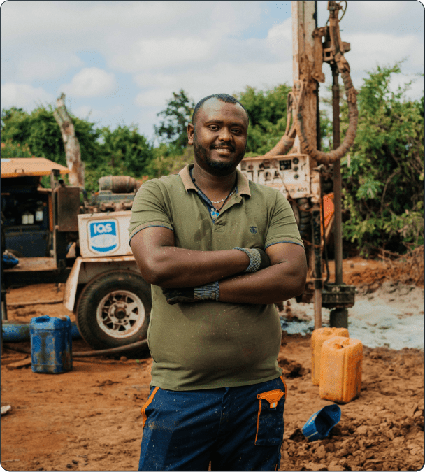 A man in a green shirt and gloves stands with arms crossed at a drilling site, with machinery, water containers, and trees in the background on a cloudy day.
