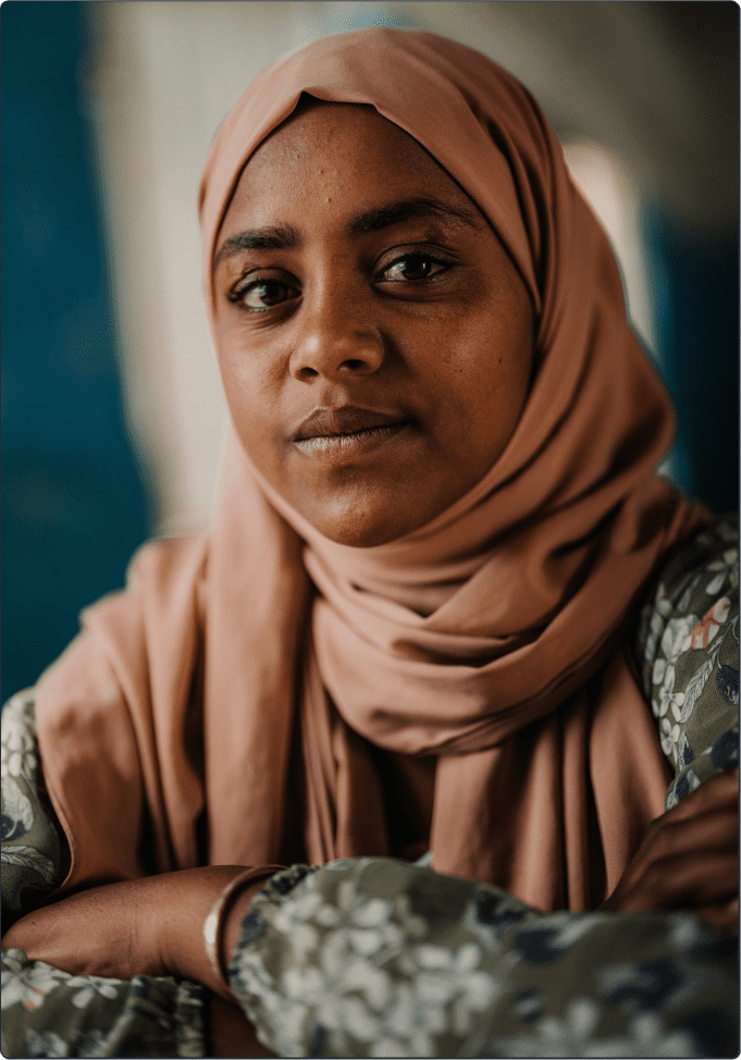 A woman wearing a light brown hijab and a floral-patterned top looks calmly at the camera with her arms crossed. The background is softly blurred in shades of blue and beige.