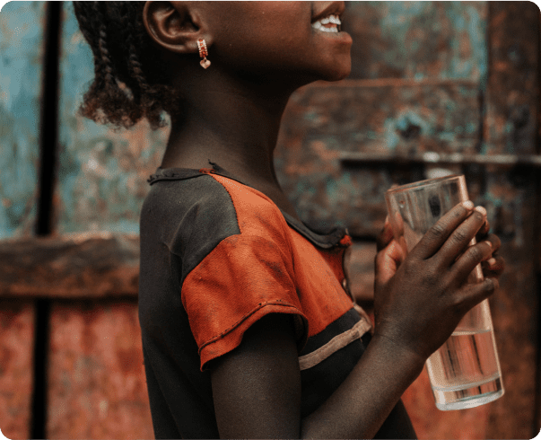 A young child smiles while holding a glass of clear water with both hands. The child wears a black and orange shirt and small earrings, standing in front of a weathered blue and brown door.