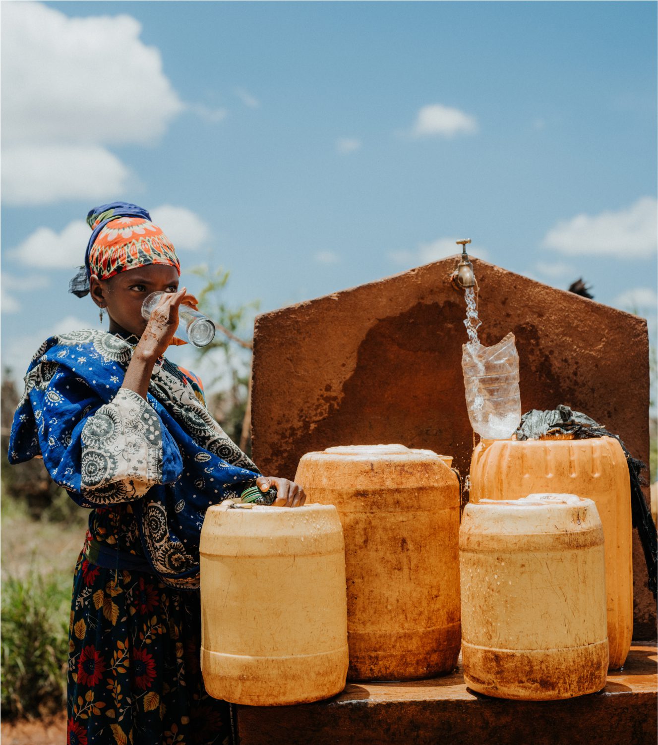 A woman in colorful clothing drinks water from a glass beside large yellow containers and a running tap outdoors under a blue sky.