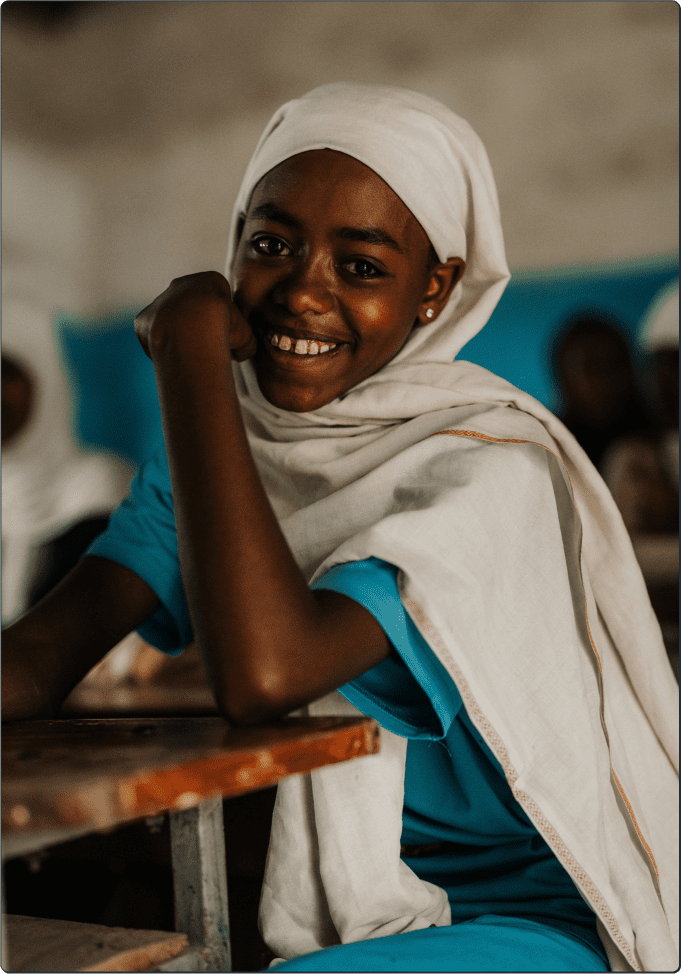 A smiling girl wearing a white headscarf and a blue shirt sits at a wooden desk in a classroom, resting her arm on the desk and looking at the camera. Other students are blurred in the background.