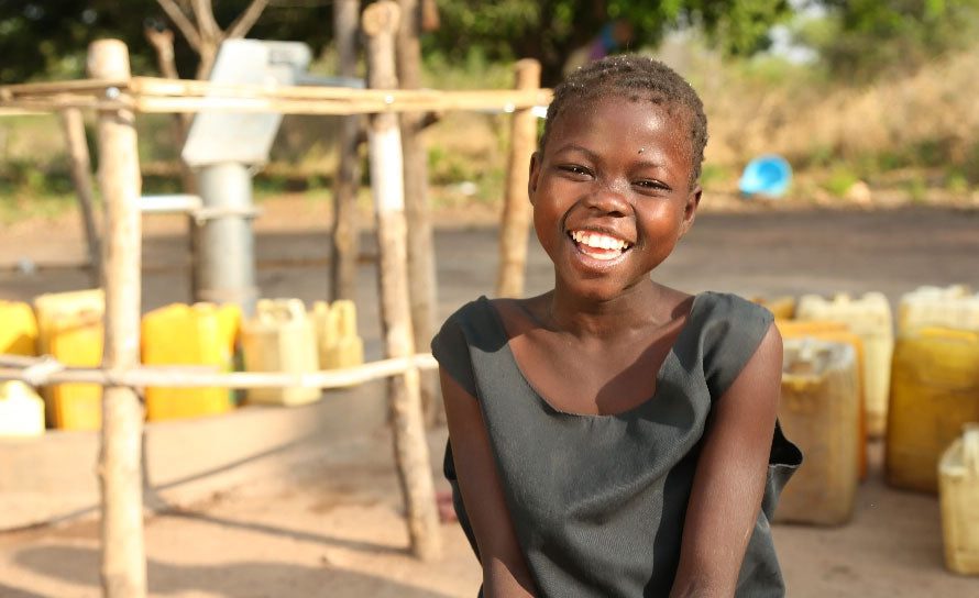 A smiling young girl in a dark dress sits outside near a water pump, surrounded by yellow jerry cans, with trees and dry grass in the background.