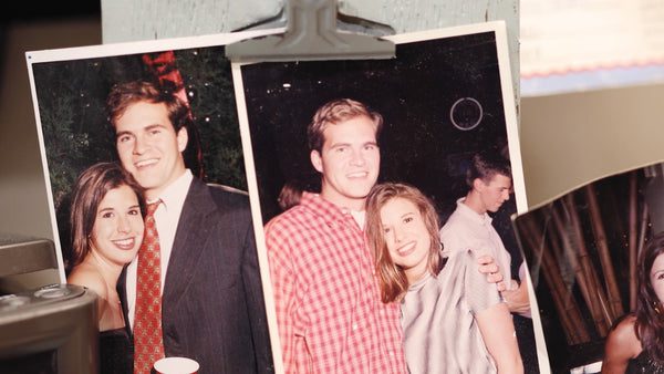 Two old photos clipped to a board show a young man and woman smiling together at social events; in one, the man wears a suit, in the other, he wears a red plaid shirt and she has her arm around him.