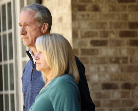 A man and woman stand side by side, looking thoughtfully into the distance outside a building with tan brick walls and large windows.