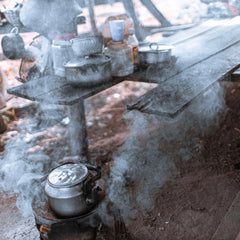 A metal kettle emits steam on a small outdoor stove, with pots, cups, and utensils scattered on a rustic wooden table nearby. Smoke and haze fill the scene, suggesting cooking in an open-air setting.