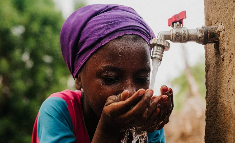 A young girl wearing a purple headscarf drinks fresh water from a tap, cupping her hands under the flowing stream on a sunny day, with a blurred green background.