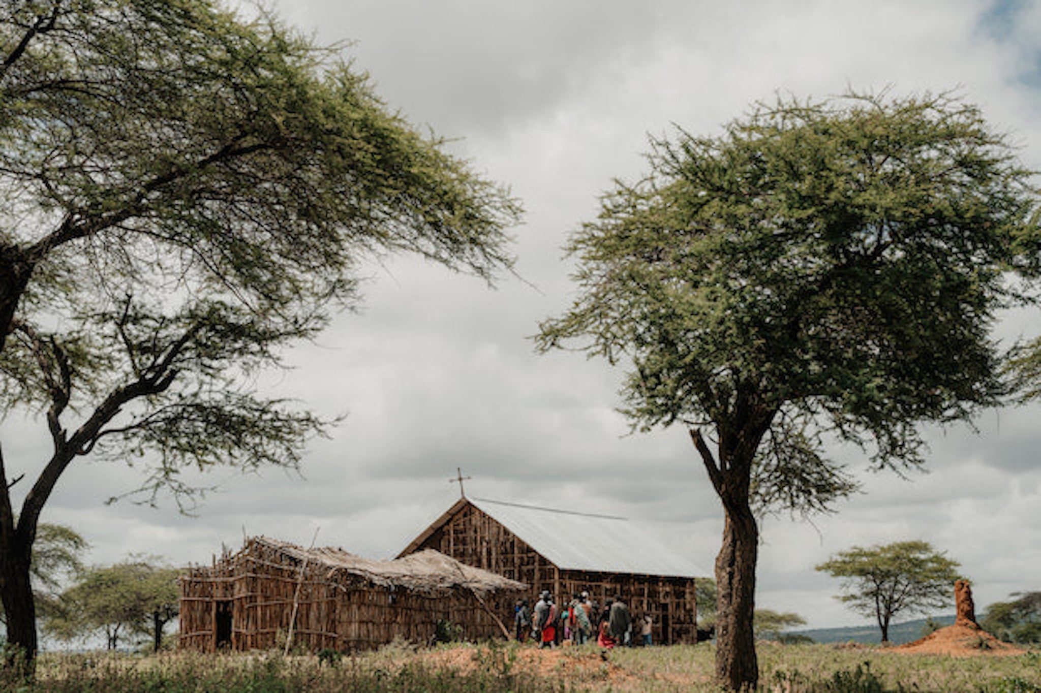 A rustic wooden church with a tin roof stands under large acacia trees. Several people gather outside on a grassy field under a cloudy sky.