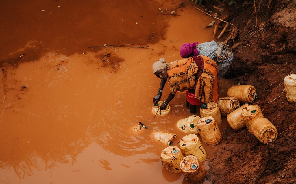 Two women fill yellow plastic containers with muddy water from a brown, polluted riverbank. The women wear colorful clothes and headscarves, and several containers are scattered around them on the ground.