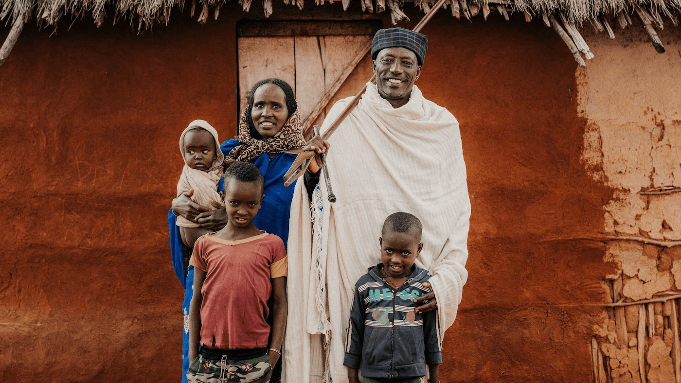 A smiling family of five stands in front of a rustic, reddish-brown mud house with a thatched roof. The parents wear traditional clothing, and the three children stand closely in front of them, smiling at the camera.