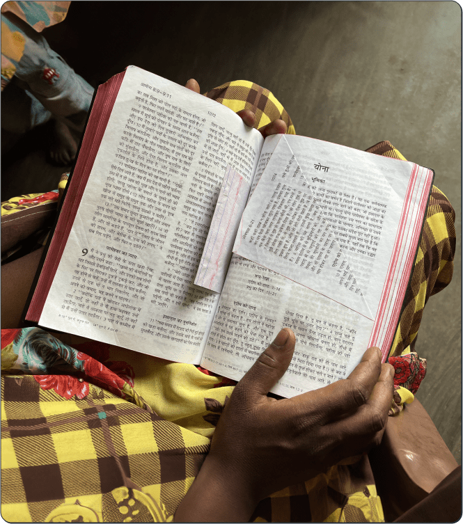 A person wearing a yellow checkered garment holds an open Hindi book in their lap, with a paper slip marking one of the pages. The setting appears indoors with soft natural light.