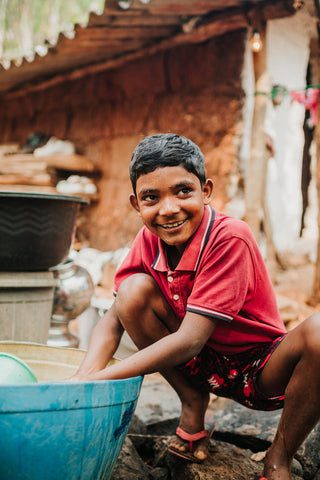 A young boy in a red shirt and shorts smiles while crouching beside a large blue basin, washing something. The background shows rustic structures and household items, suggesting a rural setting.