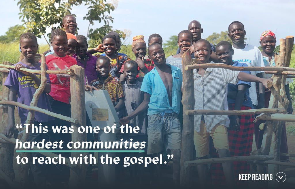 A group of smiling children and adults stand together outdoors by a wooden fence. Overlaid text reads: “This was one of the hardest communities to reach with the gospel.”.