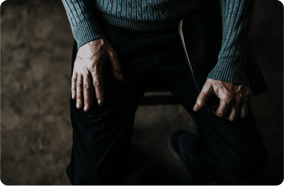 A person wearing a gray sweater and dark pants sits with both hands resting on their knees, holding a closed book in one hand. The lighting is dim, highlighting the texture of their hands and clothing.