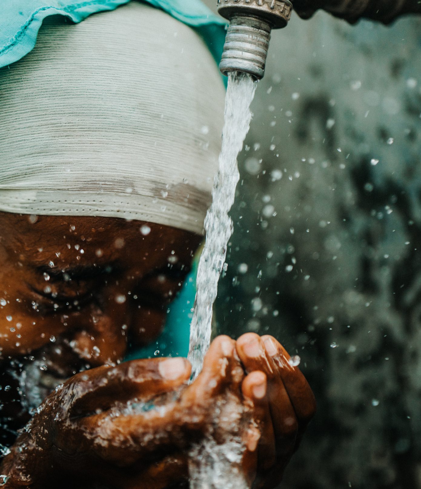 A person wearing a light-colored head covering catches and drinks water from a running tap, splashing water over their face and hands.