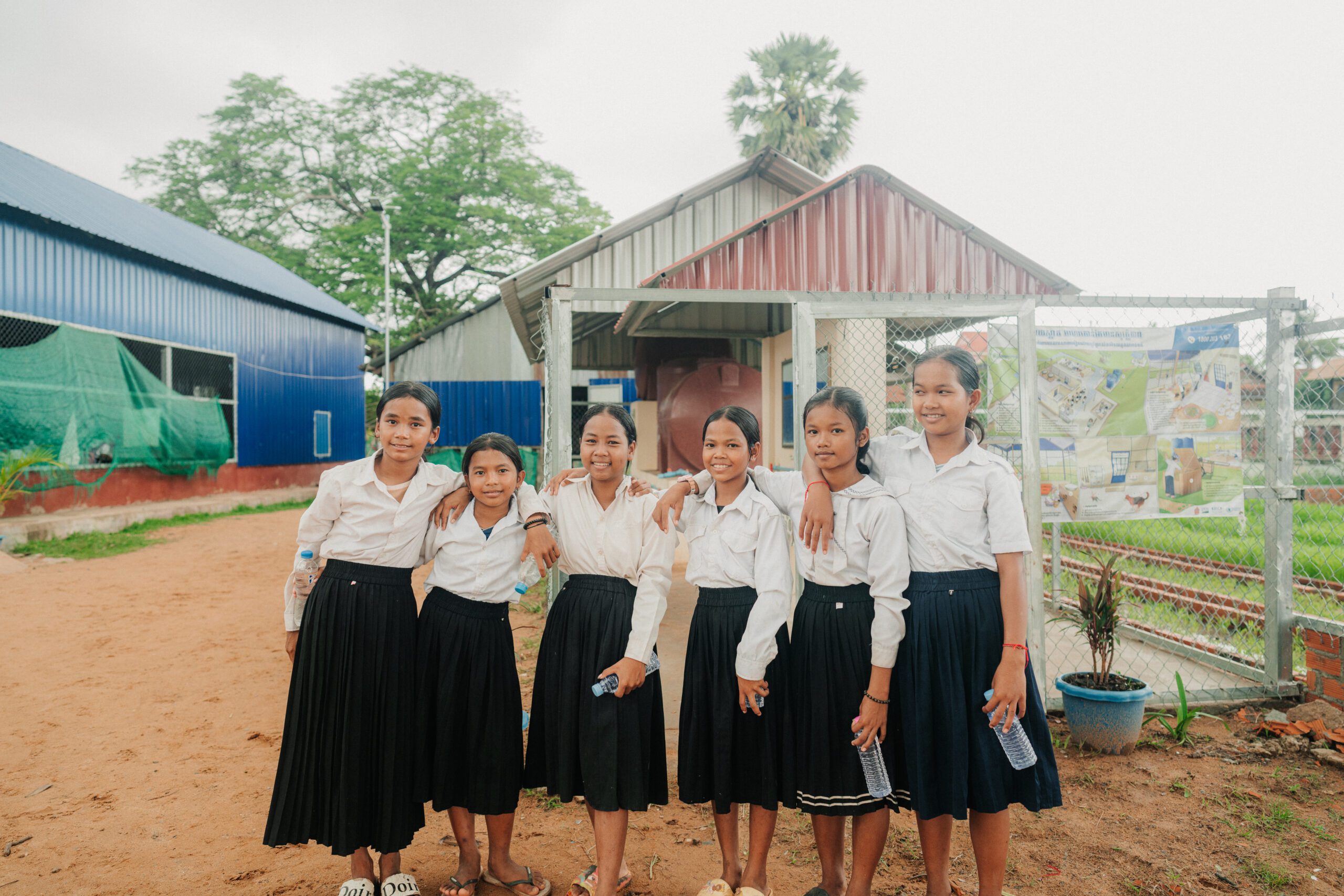 Six girls in white shirts and dark skirts stand together outside, smiling at the camera. They are holding water bottles and appear to be at a school with buildings and a fence in the background.