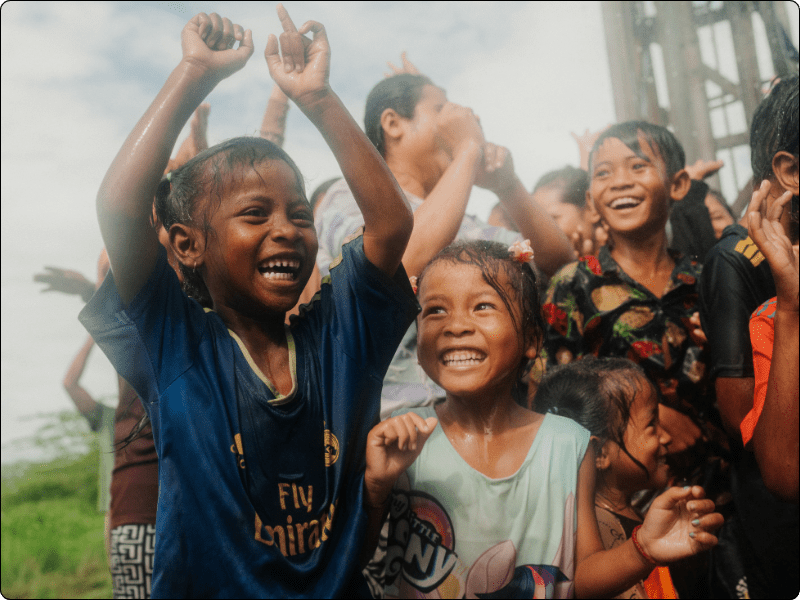 A group of joyful children, some with wet clothes and hair, smile and cheer outdoors on a cloudy day. The kids appear to be celebrating, with their hands raised and expressions of excitement and happiness.