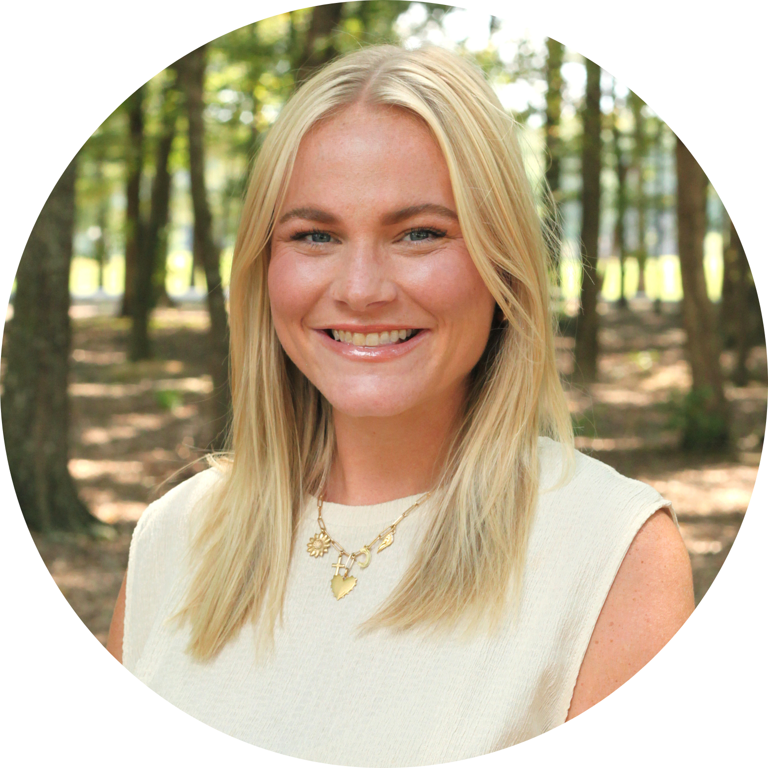 A smiling woman with straight blonde hair wearing a cream sleeveless top and a gold necklace, standing outdoors in a sunlit forested area.