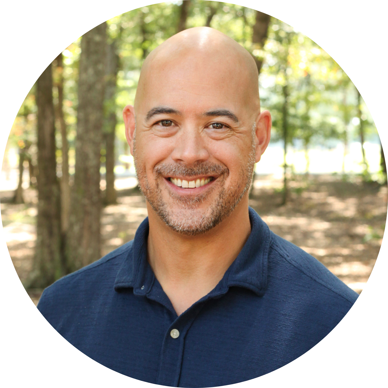 A smiling man with a shaved head, trimmed beard, and wearing a navy blue polo shirt stands outdoors in a wooded area with sunlight filtering through the trees.