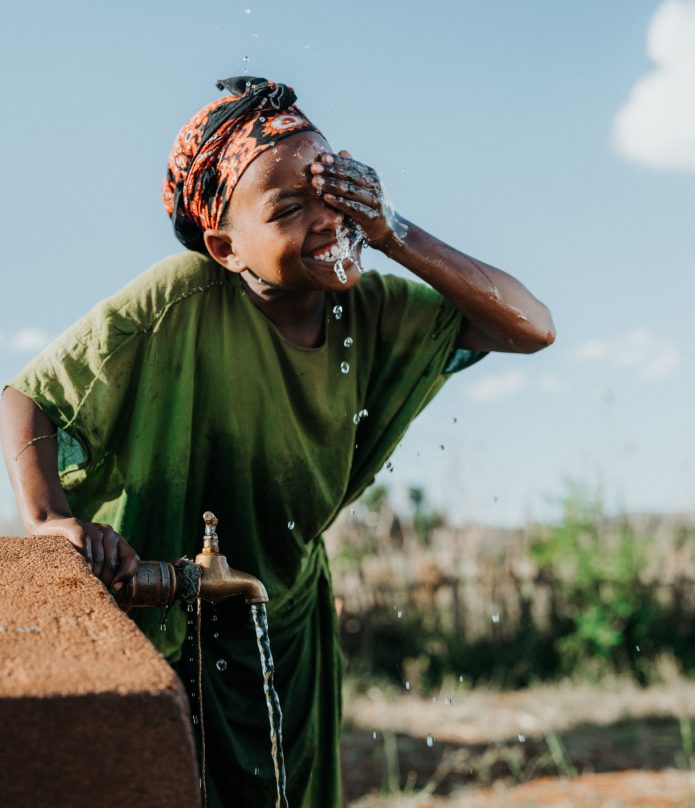 Rectangle 2 A smiling child in a green shirt splashes water on their face from an outdoor tap, with water droplets visible in the air and greenery in the background under a clear sky.