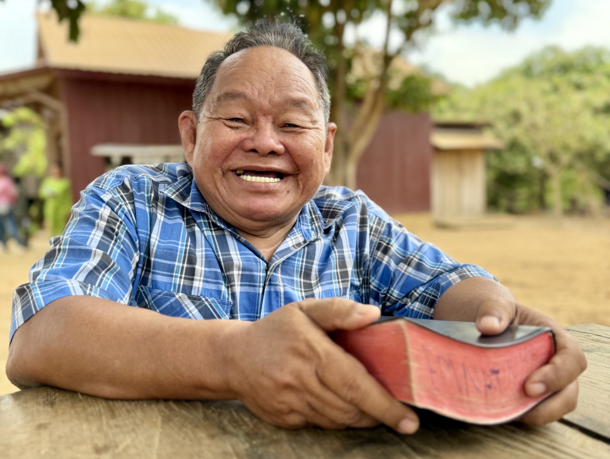 Smiling man in a blue plaid shirt sits at a wooden table outdoors, holding a thick book with red-edged pages. Trees and rustic buildings are visible in the background on a sunny day.