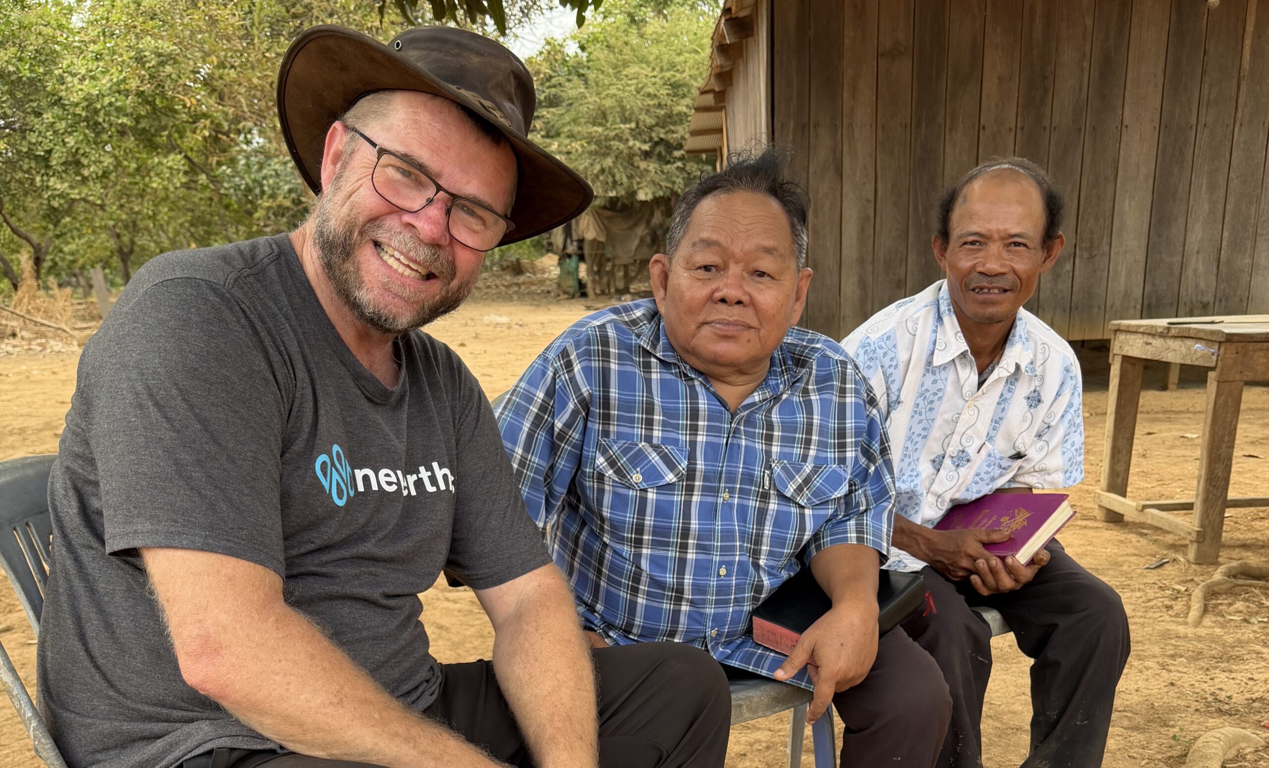 Three men sit outdoors in front of a wooden building. One wears a hat and glasses, smiling, while the other two hold books and look at the camera. Trees and dry ground are visible in the background.